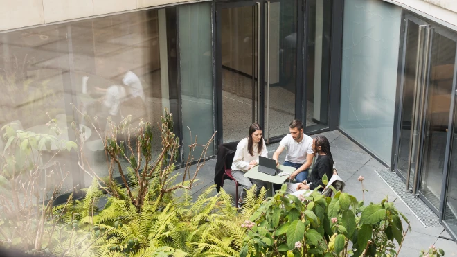 Two women, one man sit at a small outdoor table, looking at a laptop. Green foliage is in the foreground, large glass doors behind.